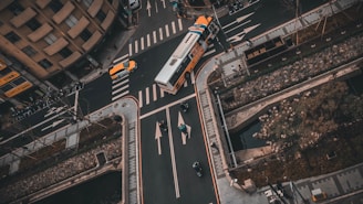 An aerial view of a city street intersection with vehicles including a bus and a taxi navigating through the lanes. Crosswalks and road markings are visible, as well as some pedestrians and cyclists. Surrounding the intersection are buildings, sidewalks, and landscaped areas with trees and shrubs.