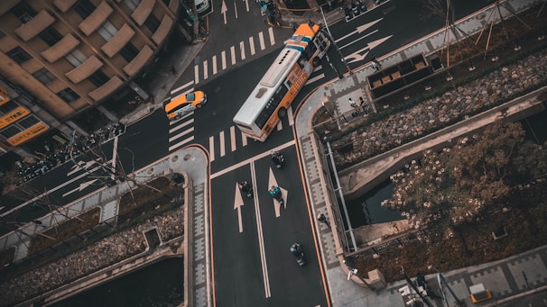An aerial view of a city street intersection with vehicles including a bus and a taxi navigating through the lanes. Crosswalks and road markings are visible, as well as some pedestrians and cyclists. Surrounding the intersection are buildings, sidewalks, and landscaped areas with trees and shrubs.