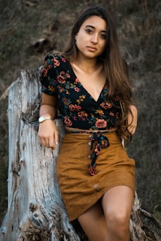 A woman with long hair is leaning against a weathered tree stump. She is wearing a floral patterned black top and a brown skirt, accessorized with a wristwatch on her left wrist. The background consists of natural, earthy textures.