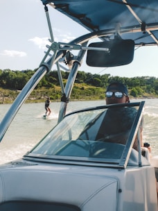 A person driving a speedboat on a lake with a wakeboarder being towed behind. The driver is wearing sunglasses and a cap, while the wakeboarder is visible in the background against a backdrop of green trees and a clear sky.