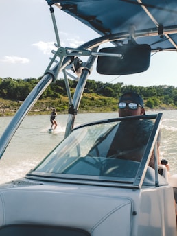 A person driving a speedboat on a lake with a wakeboarder being towed behind. The driver is wearing sunglasses and a cap, while the wakeboarder is visible in the background against a backdrop of green trees and a clear sky.