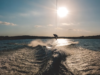 A person is performing a mid-air trick while wakeboarding on a large body of water. The sun is low in the sky, casting a warm glow over the scene, and there are waves trailing behind a boat. The silhouette of the person and their board is highlighted against the bright sunlight.