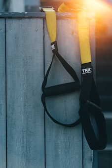 A set of TRX suspension training straps hangs from a wooden surface. The straps are predominantly black with bright yellow accents. The lighting creates a warm glow in the top right corner, adding a sunlit effect.