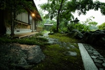 A peaceful garden scene with a stone path leading to a traditional wooden building. Lush greenery and moss cover the ground, and trees with dense foliage provide a serene atmosphere. There is a stone lantern visible in the background, adding to the tranquil and natural setting. Soft lighting from the house enhances the calm mood.