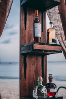 Several bottles of liquor are displayed on a wooden shelf outdoors, with a beach and ocean visible in the background. The scene is framed by fabric curtains, creating a relaxed and rustic atmosphere.