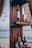 Several bottles of liquor are displayed on a wooden shelf outdoors, with a beach and ocean visible in the background. The scene is framed by fabric curtains, creating a relaxed and rustic atmosphere.