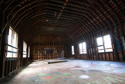A spacious, empty wooden room with a curved ceiling framework. The floor has scattered colorful graffiti and debris. Natural light streams through the large, rectangular windows lining the walls.