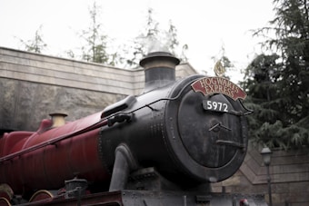 A vintage steam train engine is positioned outdoors, prominently displaying a sign that reads 'Hogwarts Express' and the number '5972'. The train is deep red and black, with metal rivets and pipes visible, suggesting a classic design. Behind the train, there are stone walls and trees, enhancing the rustic and historic atmosphere.