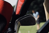 A close-up view of a set of golf clubs is prominently featured, with a blurred background showing a person playing golf on a green field. The focus is on the metal clubs with red and black head covers, while the golfer appears out of focus in the background.