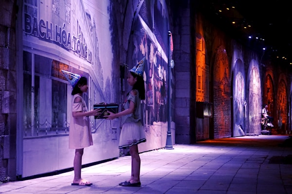 Two children wearing party hats are standing on a dimly lit street, facing each other. They appear to be exchanging a gift. The background features large, illuminated murals along a wall, creating a colorful and festive atmosphere.