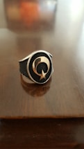 A metallic ring with a textured black background features a prominent crescent and star design in the center. The background is a smooth, polished wooden surface, enhancing the stark contrast of the ring's shiny metal and dark inlay.