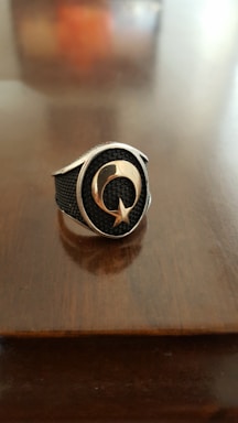 A metallic ring with a textured black background features a prominent crescent and star design in the center. The background is a smooth, polished wooden surface, enhancing the stark contrast of the ring's shiny metal and dark inlay.