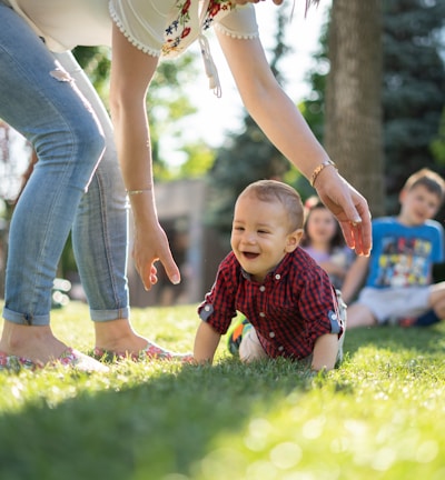 woman in white blouse and blue denim jeans helping a baby crawl on green grass