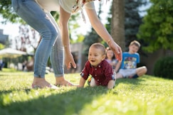 woman in white blouse and blue denim jeans helping a baby crawl on green grass