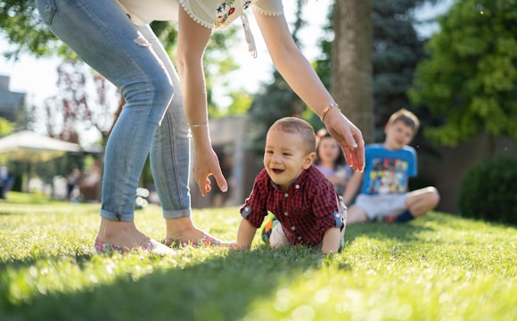 woman in white blouse and blue denim jeans helping a baby crawl on green grass
