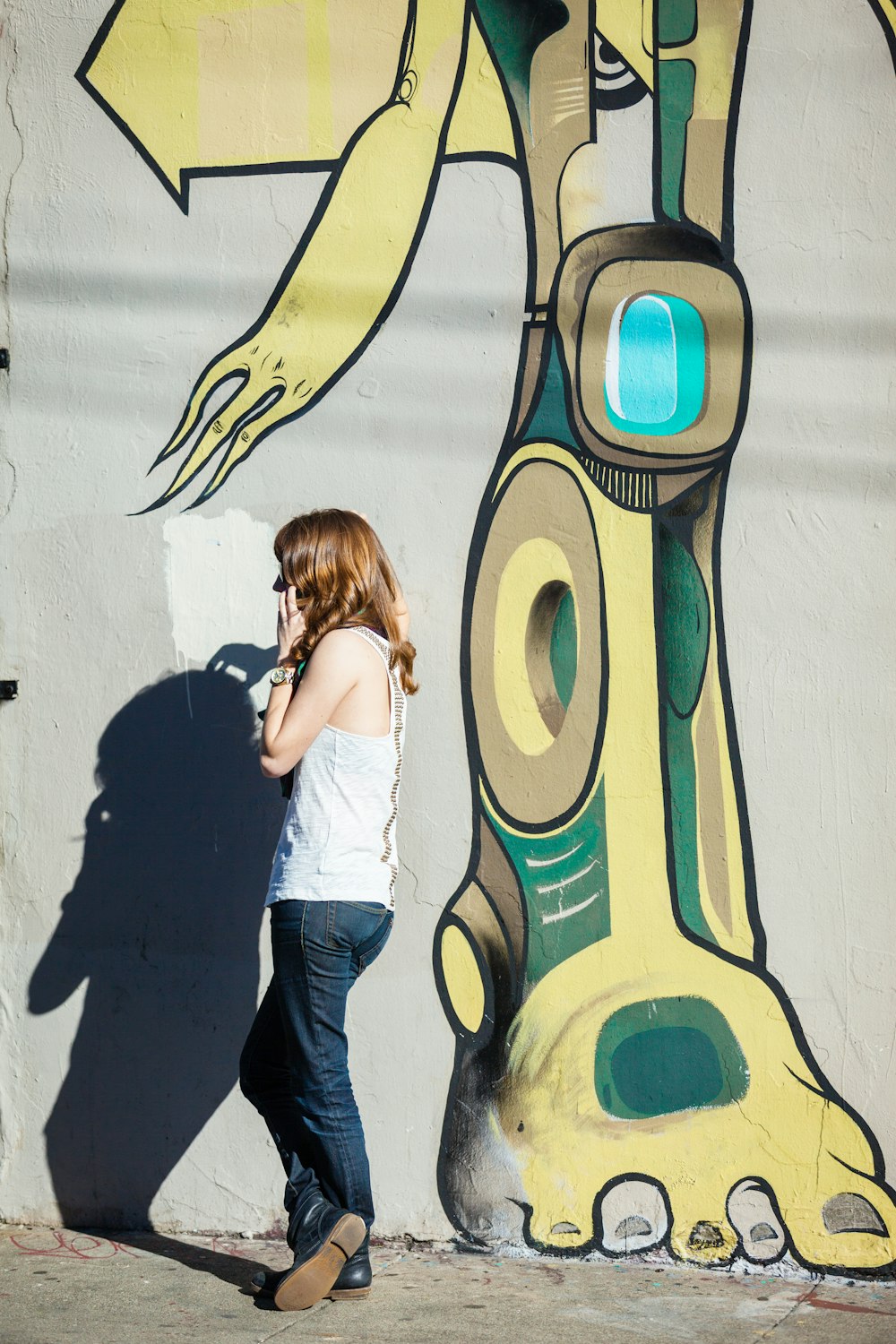 woman in white sleeveless shirt and blue denim jeans stands beside a grey wall with yellow and green robot graffiti