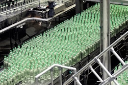 A production line filled with rows of clear plastic bottles capped with green lids. The bottles are neatly organized and moving along a conveyor in an industrial setting, indicating a bottling plant or factory environment.