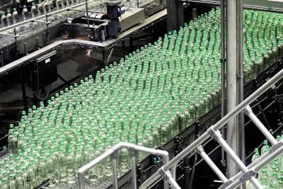 A production line filled with rows of clear plastic bottles capped with green lids. The bottles are neatly organized and moving along a conveyor in an industrial setting, indicating a bottling plant or factory environment.