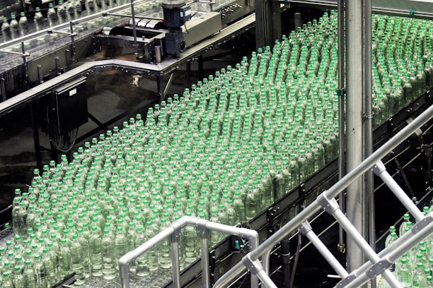 A production line filled with rows of clear plastic bottles capped with green lids. The bottles are neatly organized and moving along a conveyor in an industrial setting, indicating a bottling plant or factory environment.