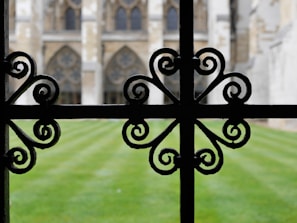 An ornate black wrought iron fence with intricate, symmetrical scrollwork patterns is in focus in the foreground. Beyond the fence, a well-maintained lawn leads to a historic structure featuring large arched windows and intricate stonework.