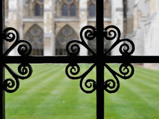An ornate black wrought iron fence with intricate, symmetrical scrollwork patterns is in focus in the foreground. Beyond the fence, a well-maintained lawn leads to a historic structure featuring large arched windows and intricate stonework.