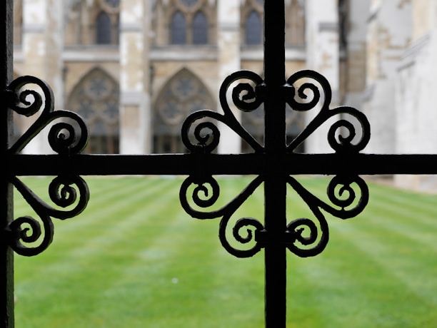 An ornate black wrought iron fence with intricate, symmetrical scrollwork patterns is in focus in the foreground. Beyond the fence, a well-maintained lawn leads to a historic structure featuring large arched windows and intricate stonework.