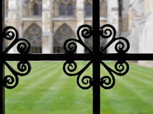 An ornate black wrought iron fence with intricate, symmetrical scrollwork patterns is in focus in the foreground. Beyond the fence, a well-maintained lawn leads to a historic structure featuring large arched windows and intricate stonework.