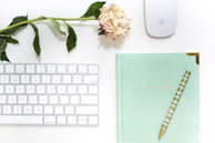 A minimalist workspace featuring a white keyboard, a wireless mouse, and a mint green notebook with gold corner details. A striped pen rests diagonally across the notebook, which has 'FULL OF IDEAS' embossed in gold. A single pink peony with green leaves is placed above the keyboard, adding a touch of natural elegance.
