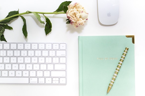 A minimalist workspace featuring a white keyboard, a wireless mouse, and a mint green notebook with gold corner details. A striped pen rests diagonally across the notebook, which has 'FULL OF IDEAS' embossed in gold. A single pink peony with green leaves is placed above the keyboard, adding a touch of natural elegance.