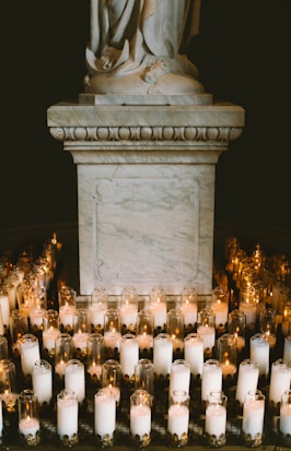 A marble statue pedestal surrounded by numerous lit candles in glass holders, creating a serene and contemplative atmosphere. The soft glow from the candles casts gentle light and shadows on the statue's base.
