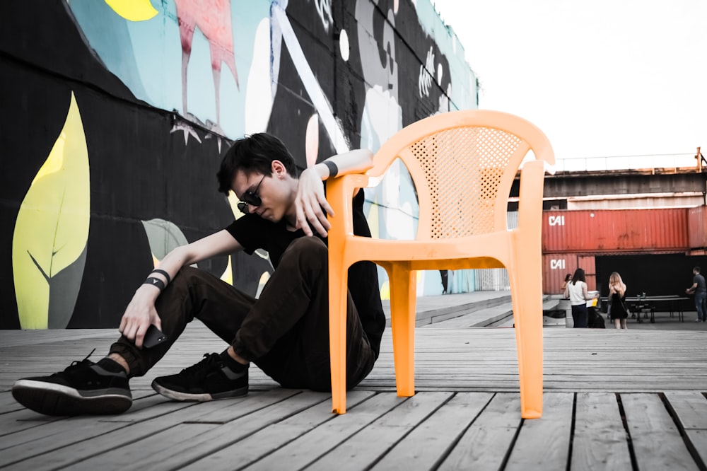 man sitting on floor beside empty orange plastic monobloc armchair at daytime