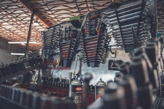 A workshop scene features a well-organized wall with an array of tools including wrenches, sockets, and screwdrivers. The tools are neatly arranged and displayed on a metallic surface, with a workbench beneath holding cans of spray and other items. Overhead, beams of the workshop ceiling are visible, providing an industrial backdrop.