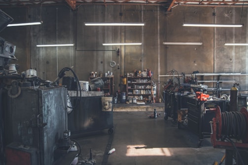 A dimly lit garage or workshop space filled with industrial equipment and tools. Metal shelves are stocked with various containers and boxes, while several large machines are positioned around the room. The concrete walls and floor contribute to a gritty and worn atmosphere. Overhead fluorescent lights cast a harsh glow, adding to the utilitarian feel of the space.