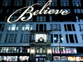 A large building facade illuminated with a festive sign reading 'Believe' in bright lights. The architecture is classic with multiple windows, and the entrance is adorned with statues and a clock. There are street lamps and storefronts at the street level with colorful displays and a small gathering of people.