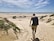 A person with a backpack walks barefoot on sandy dunes towards an ocean. In the background, there are several vehicles parked along a path parallel to the shoreline. The sky is clear with a few clouds, and some greenery is visible on the sides of the dunes.