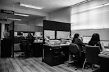 A monochrome office environment with several people working at desks. Stacks of papers and computer screens are visible, creating a busy workplace setting. The office is divided by low partitions, and there are fluorescent lights on the ceiling.