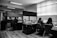 A monochrome office environment with several people working at desks. Stacks of papers and computer screens are visible, creating a busy workplace setting. The office is divided by low partitions, and there are fluorescent lights on the ceiling.