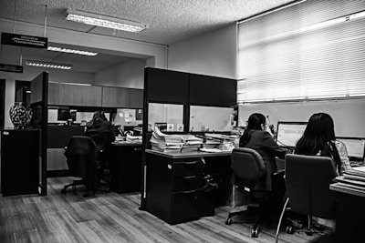 A monochrome office environment with several people working at desks. Stacks of papers and computer screens are visible, creating a busy workplace setting. The office is divided by low partitions, and there are fluorescent lights on the ceiling.