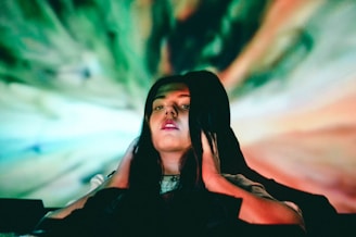 A person with long dark hair holds their hands to their head, immersed in a colorful, swirling background of green, blue, and orange hues. The lighting creates a surreal and enigmatic atmosphere.