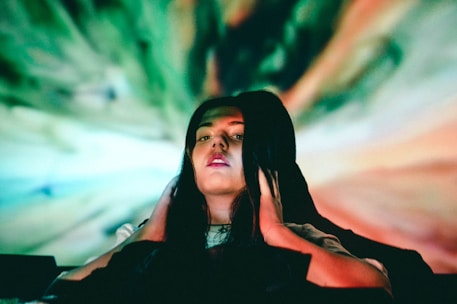 A person with long dark hair holds their hands to their head, immersed in a colorful, swirling background of green, blue, and orange hues. The lighting creates a surreal and enigmatic atmosphere.