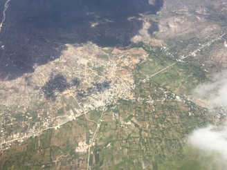 An aerial view of a landscape with a mix of urban and rural areas. The image shows a patchwork of agricultural fields, roads, and a densely built area possibly representing a small town or city. Dark shadows from clouds are cast over part of the landscape, creating a contrast with the areas illuminated by sunlight.