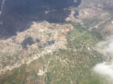 An aerial view of a landscape with a mix of urban and rural areas. The image shows a patchwork of agricultural fields, roads, and a densely built area possibly representing a small town or city. Dark shadows from clouds are cast over part of the landscape, creating a contrast with the areas illuminated by sunlight.