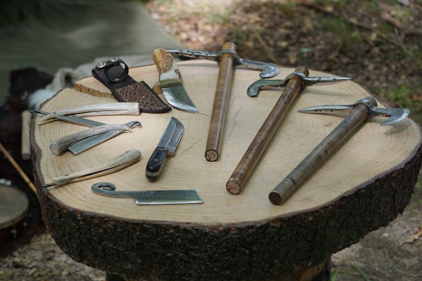 A rustic wooden display showcasing a variety of sharp bowie knives, chef knives, and Viking axes arranged with natural outdoor elements.