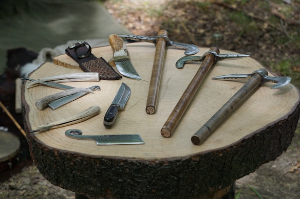 A collection of various tools and knives is neatly arranged on a wooden slab, possibly outdoors. The tools appear to have wooden or handle grips and metallic blades or components. The setting suggests an outdoor environment with natural lighting.