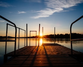 A serene lakeside scene with a wooden dock extending into the water, framed by metal railings. The sun is setting in the background, casting warm colors across the sky and water. Trees line the horizon, creating a peaceful, natural setting.