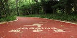 A winding red jogging path runs through a lush, green forest area. The path has white text and symbols painted on its surface, indicating it's designated for jogging. Trees and bushes line the path, providing a sense of enclosure and natural serenity.