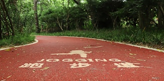 A winding red jogging path runs through a lush, green forest area. The path has white text and symbols painted on its surface, indicating it's designated for jogging. Trees and bushes line the path, providing a sense of enclosure and natural serenity.