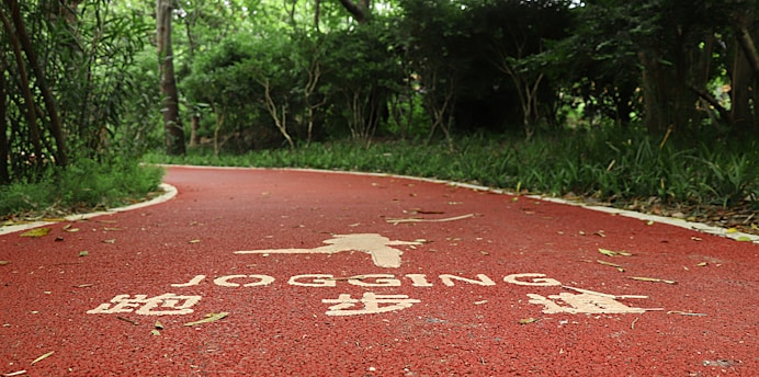 A winding red jogging path runs through a lush, green forest area. The path has white text and symbols painted on its surface, indicating it's designated for jogging. Trees and bushes line the path, providing a sense of enclosure and natural serenity.