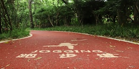 A winding red jogging path runs through a lush, green forest area. The path has white text and symbols painted on its surface, indicating it's designated for jogging. Trees and bushes line the path, providing a sense of enclosure and natural serenity.