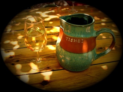 A rustic ceramic pitcher labeled 'Tishbi' sits on a wooden table alongside a glass of white wine. Sunlight filters through leaves, creating dappled patterns on the table.
